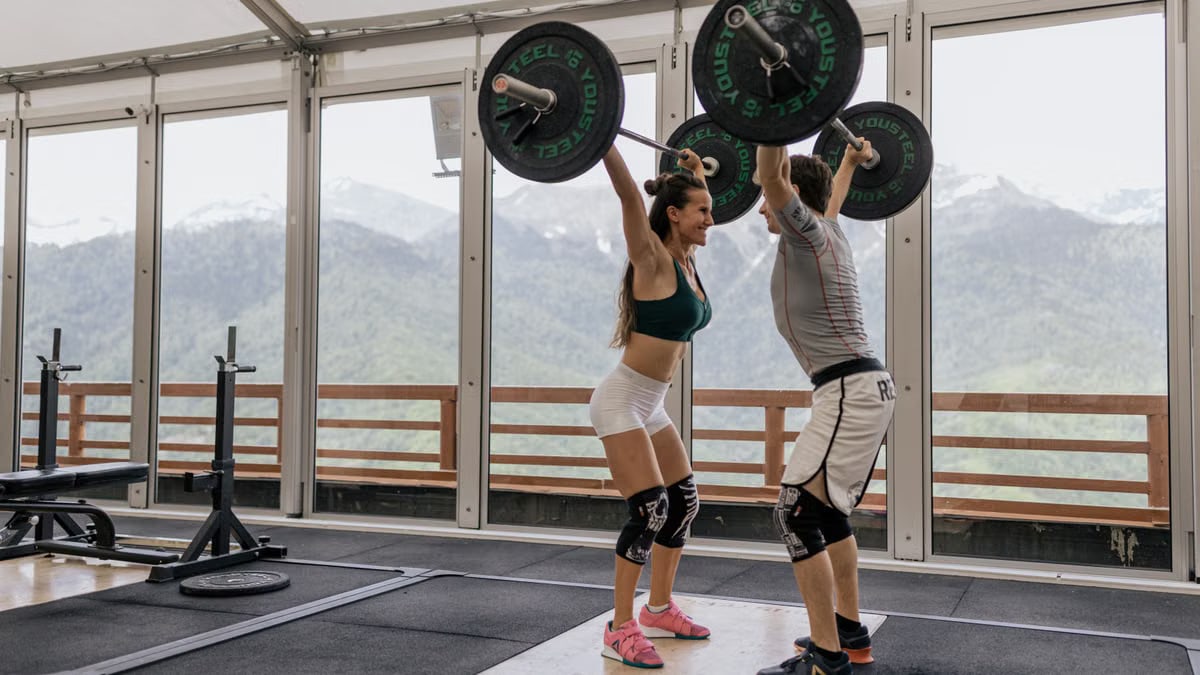 A couple lifting barbells together in a gym with scenic mountain views.